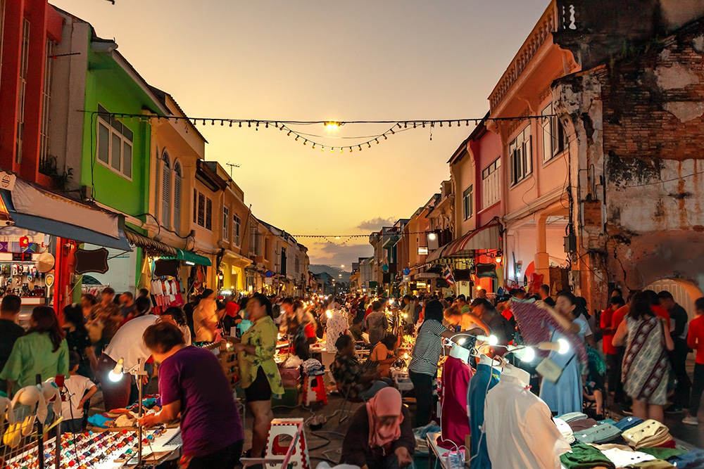 Una calle amplia con mucha gente y varios puestos de venta callejeros. Un atardecer se ve al fondo de la calle 