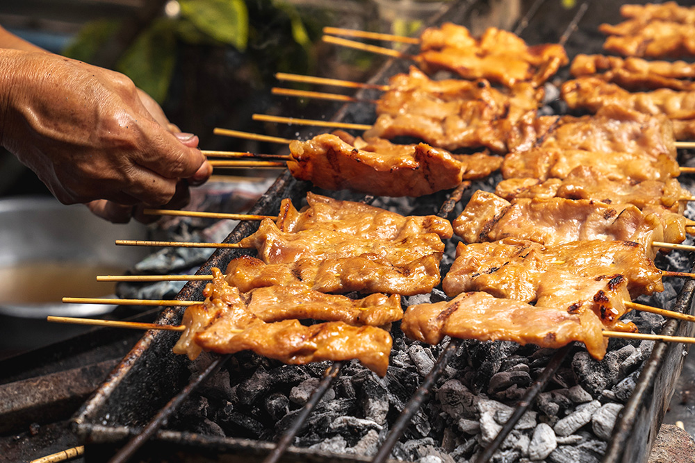 Una parrilla con carbón y encima pinchos de carne. Una mano de hombre aparece por la izquierda dando la vuelta a un pincho.