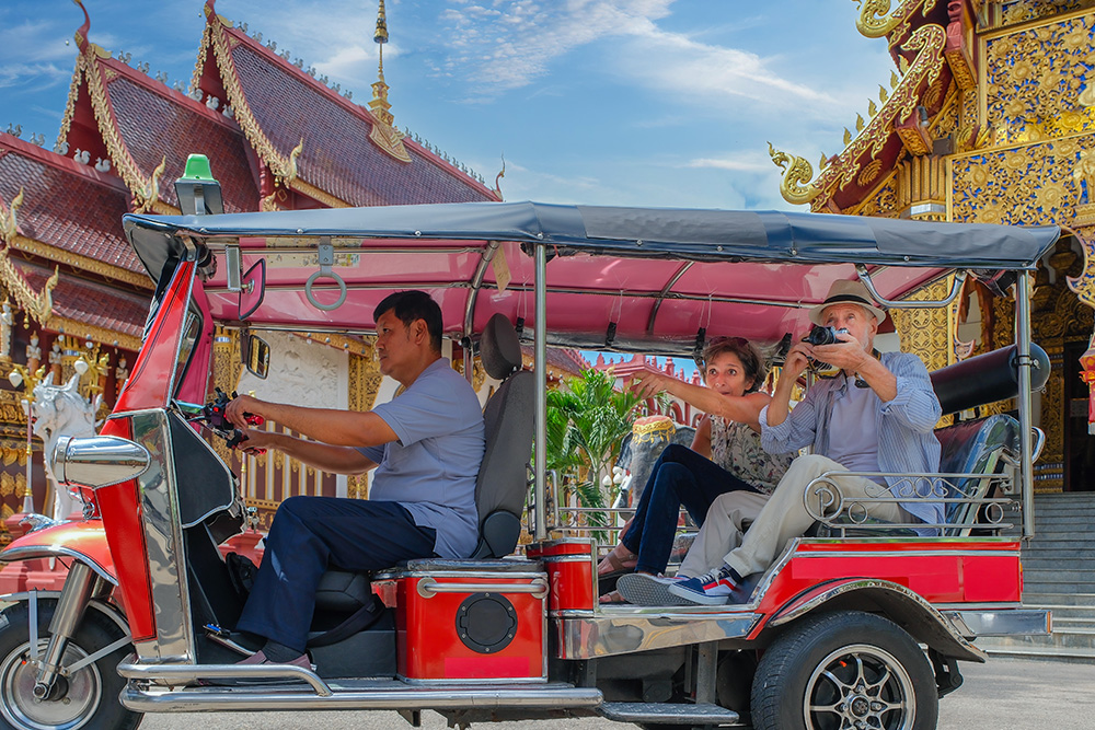Primer plano de un vehículo típico tailandés, llamado tuk-tuk con un conductor y una pareja de turistas detrás haciendo una foto