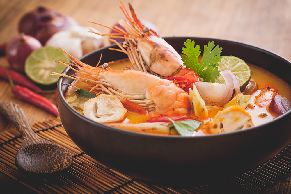 Bol con un caldo naranja y sobresaliendo unas gambas con perejil decorando y lima. Al lado del bol una cuchara de madera