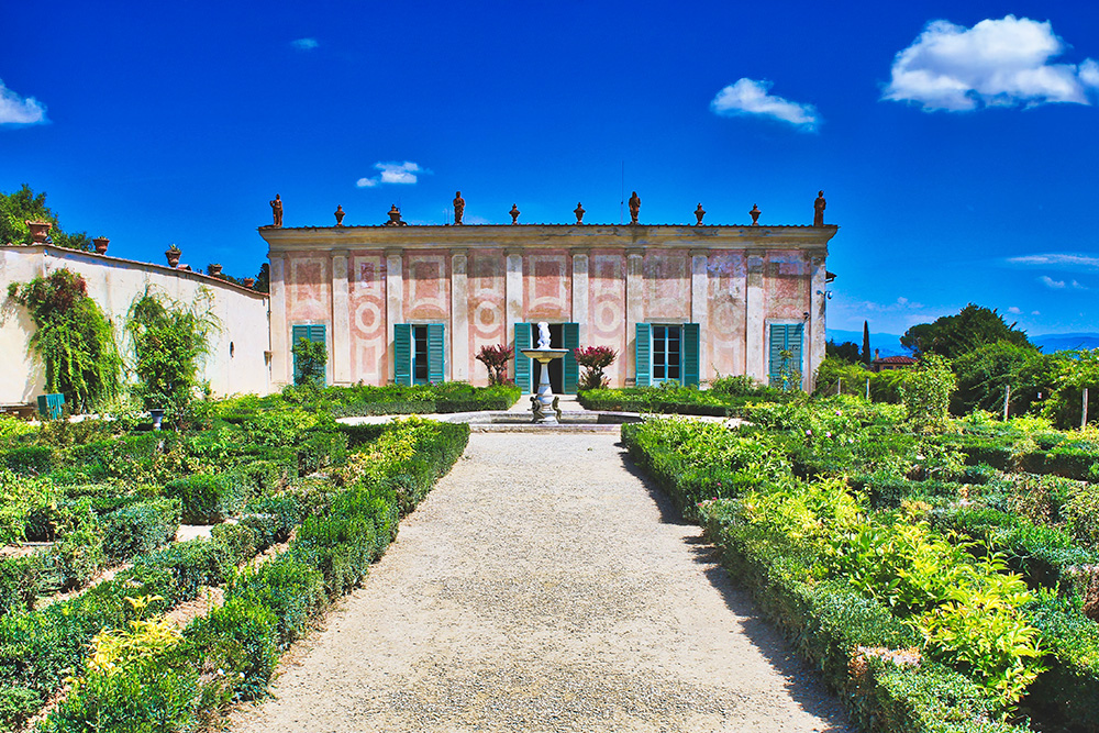 Casa rodeada de un frondoso jardin con diversos setos y caminos de tierra. En el centro y frente a la casa una pequeña fuente de piedra. 
