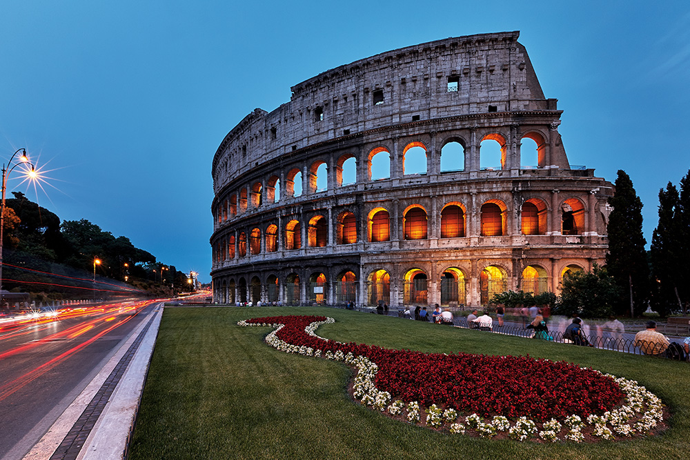 Imagen de el Coliseo en un anochecer. Alumbrado por las luces y frente a el el jardin con flores rojas y blancas.