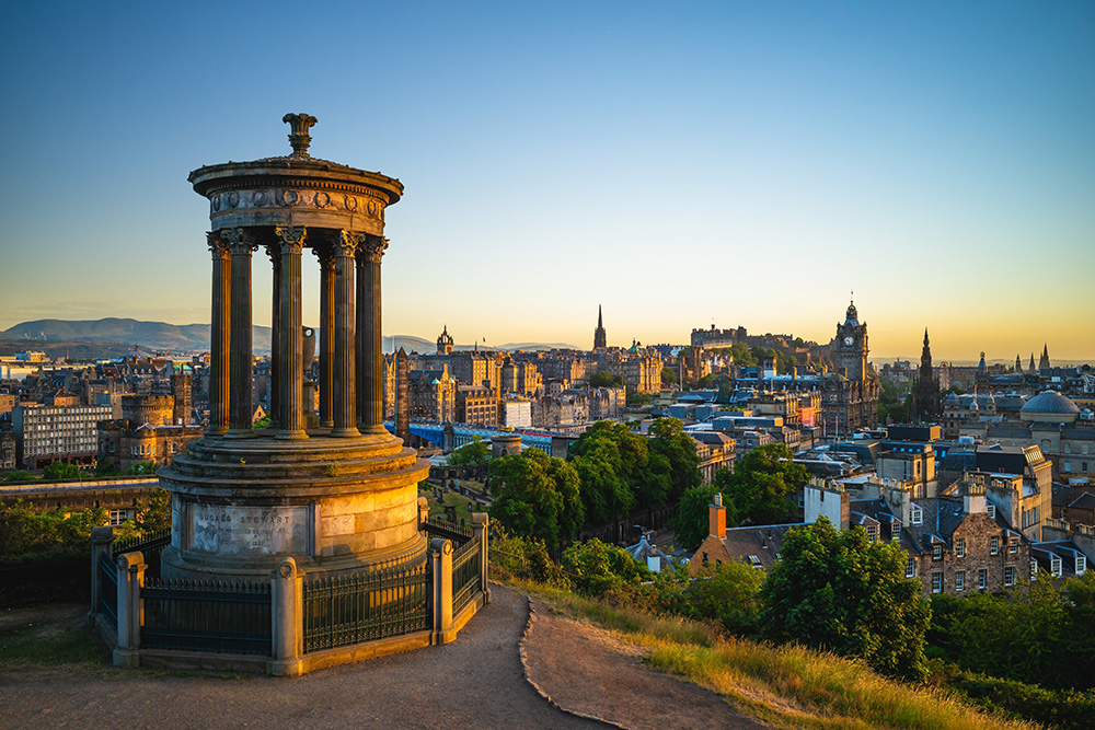 Un mirador con una torre de piedra cercada. De fondo la ciudad de Edimburgo, con sus casas de piedra y tejados acabados en punta. De fondo el cielo azul y naranja el atardecer 