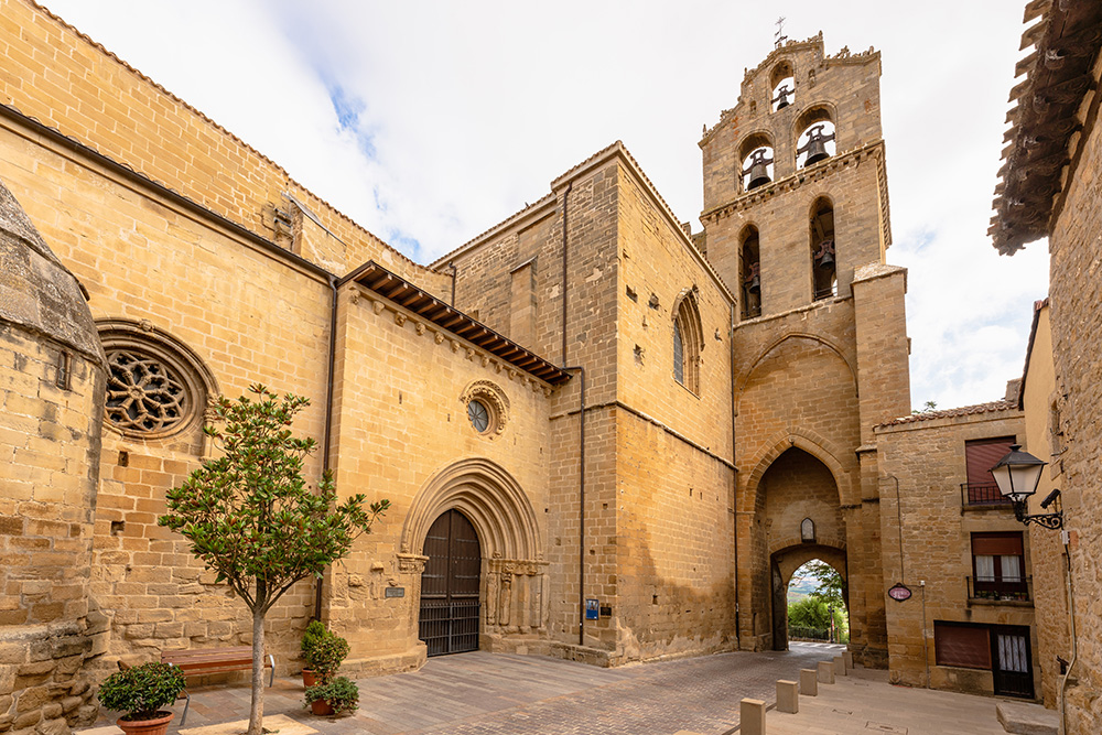 Entrada a una iglesia, con un pequeño banco a la izquierda con varias macetas y un pequeño arbol enfrente. A la derecha una entrada abovedada y el campanar encima 