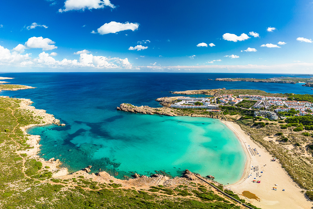 Pequeña cala de Menorca, con su mar azul turquesa. Se vislumbra el extenso mar azul marino juntándose con el cielo surcado de nubles blancas