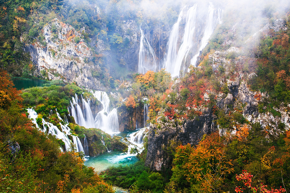 Grandes montañas llenas de vegetación verde y naranja. De las montañas surgen grandes cascadas que finalizan en un pequeño lago turquesa 