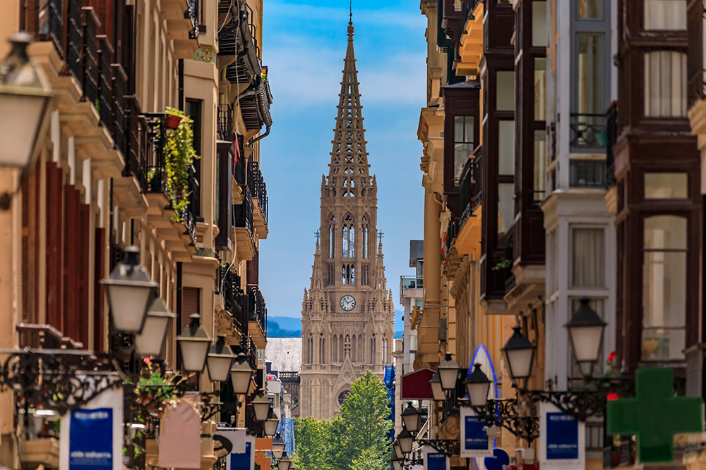 Una calle vista desde alto, con las farolas y miradores de edificios antiguos. Al fondo de la calle, el campanar de la iglesia con el reloj con un fondo de cielo azul claro. 