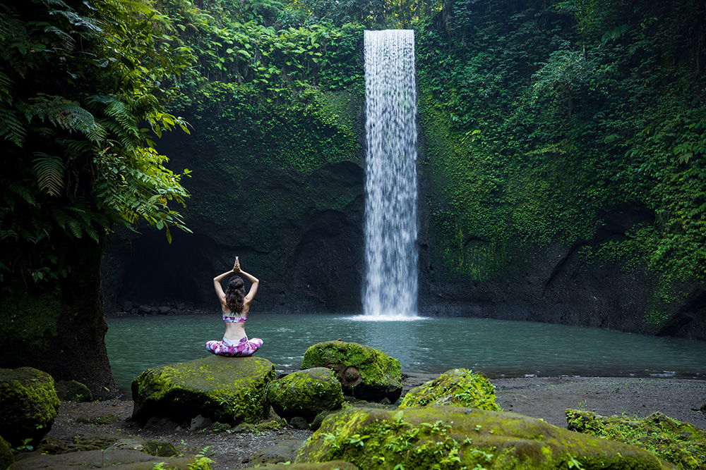 Un pequeño lago rodeado de verde y una pequeña cascada. Frente a ella una chica sentada en una roca llena de musgo verde haciendo yoga 