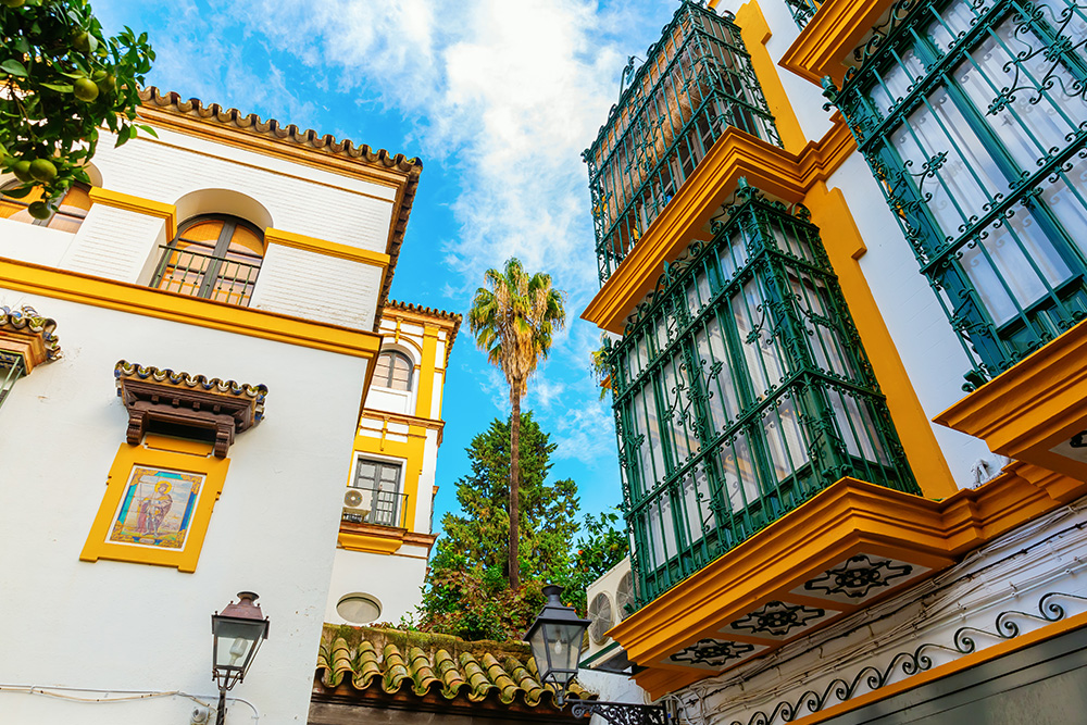 Vista hacia los balcones colgantes de una antigua casa con las rejas verdes y un cielo azul con nubes blancas de fondo 