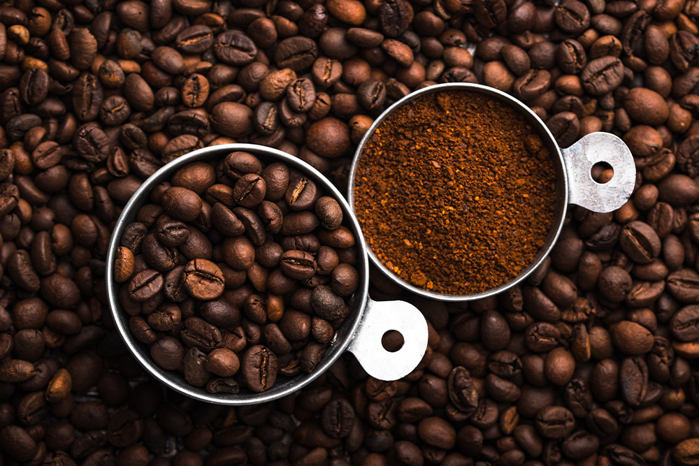 Sobre un fondo de granos de café en diversos tonos de marrón, dos cazos pequeños de metal. Uno con granos de café y el otro con café triturado.