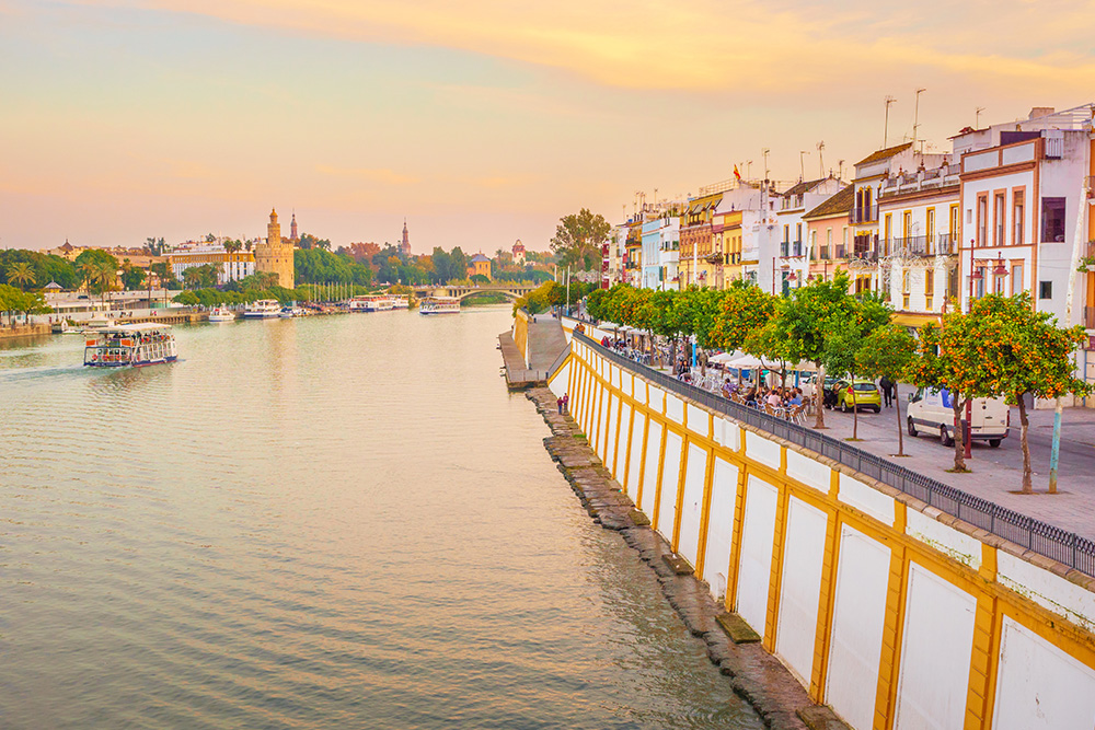 Rio Guadalquivir con una pequeña barca cruzándolo. Al lateral izquierdo las casas y el paseo que bordea el río con sus arboles 