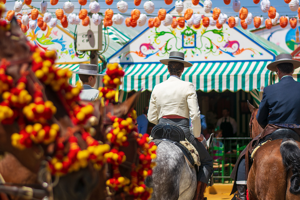 Dos hombres montando a caballo vestidos con el típo atuendo andaluz con sombrero y de fondo la entrada a una caseta andaluza con falorillos 