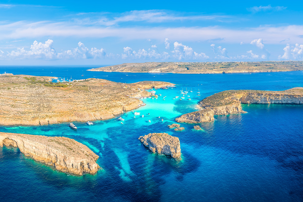 Tres islas de un tono arena en un inmenso mar de diferentes tonos de azul. Varias embarcaciones de tonos blancos en las orillas y un cielo surcado de nubes blancas