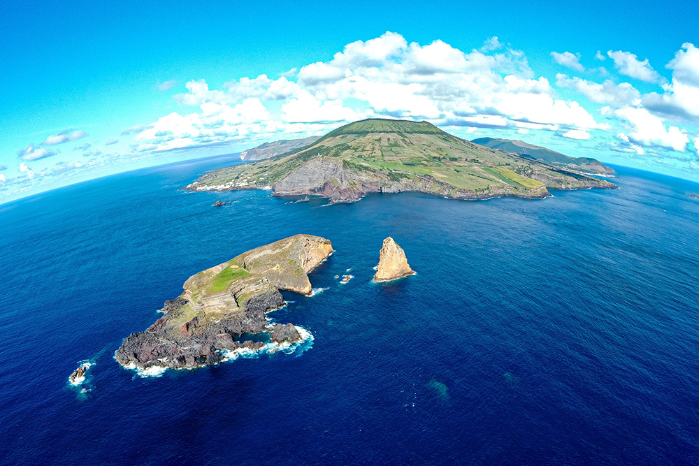 Imagen de un mar azul intenso. En el centro una pequeña isla como una montaña pequeña en tonos verdes. Frente a ella otra pequeñita isla. Cubriéndolas un cielo azul con nubes blancas