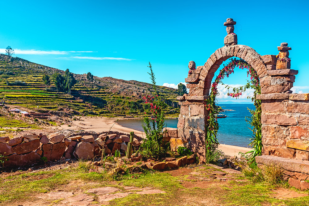 Una ladera verde con algún arbol disperso a la izquierda. A la derecha un arco de piedra con flores y tres estatuillas. Desde el arco se ve el mar y la orilla de la playa