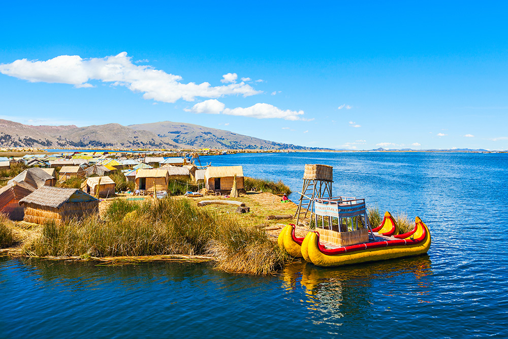 Un amplio lago de un azul marino. Se ve una pequeña parcela de tierra con casas de paja y una pequeña embarcación de dos canoas amarillas y rojas. Al fondo la montañas y nubes blancas