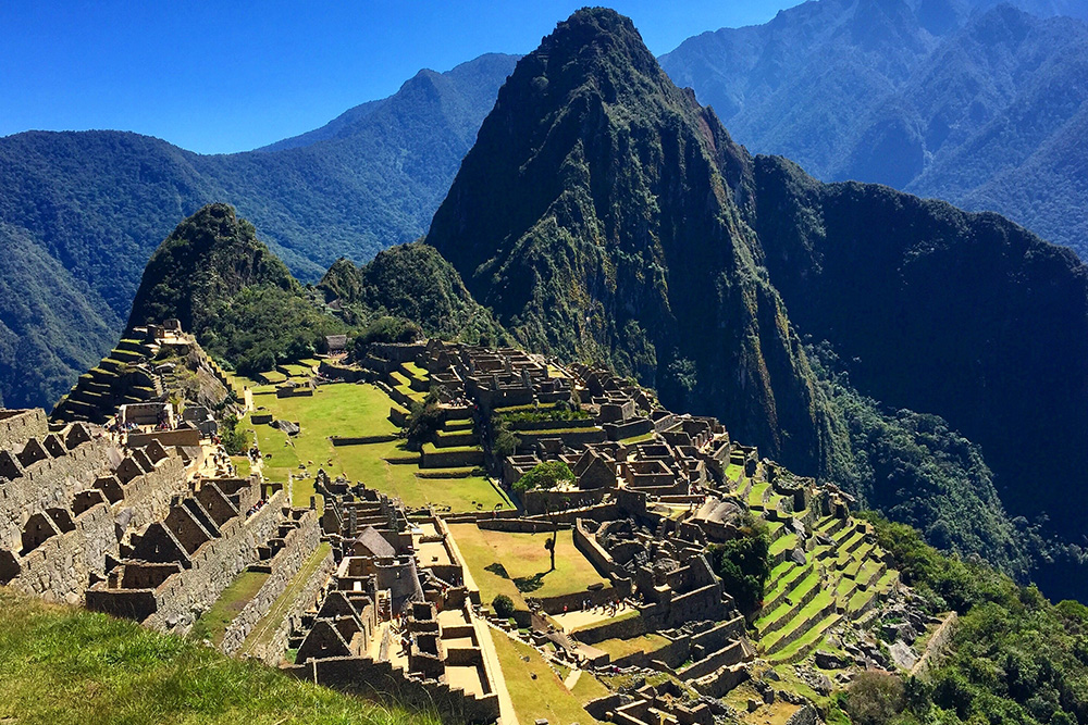 Vistas aéreas de las montañas. Se ven las cordilleras verdes al fondo enmarcadas por un cielo azul. En primer plano, un valle verde esmeralda con ruinas de piedra.