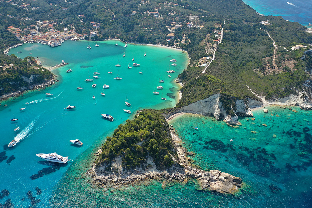 Vista aérea de una cala en forma de u con un mar azul verdoso. Se encuentra lleno de pequeñas embarcaciones. El terreno es verde y con algunas edificaciones