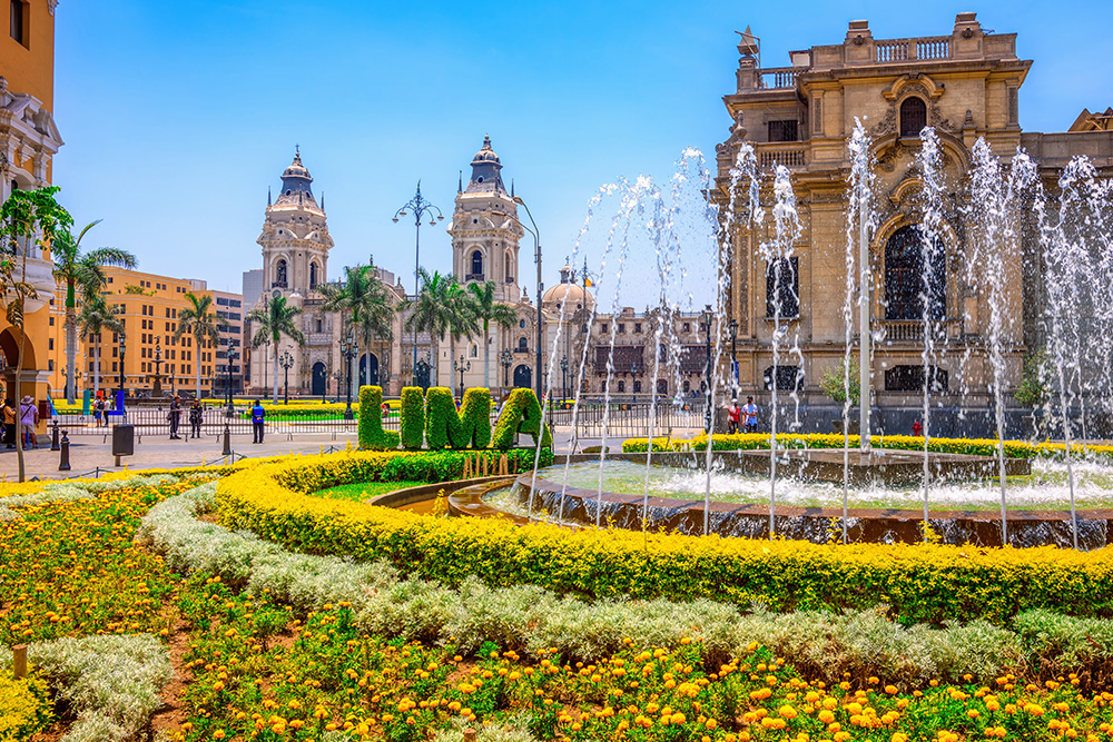 Plaza rodeada de edificios antiguos bajo un cielo azul. Palmeras frente a los edificios y en primer plano una fuente circular con con agua rodeada de flores de color amarillo y con hierba el nombre de la ciudad de Lima en un costado.