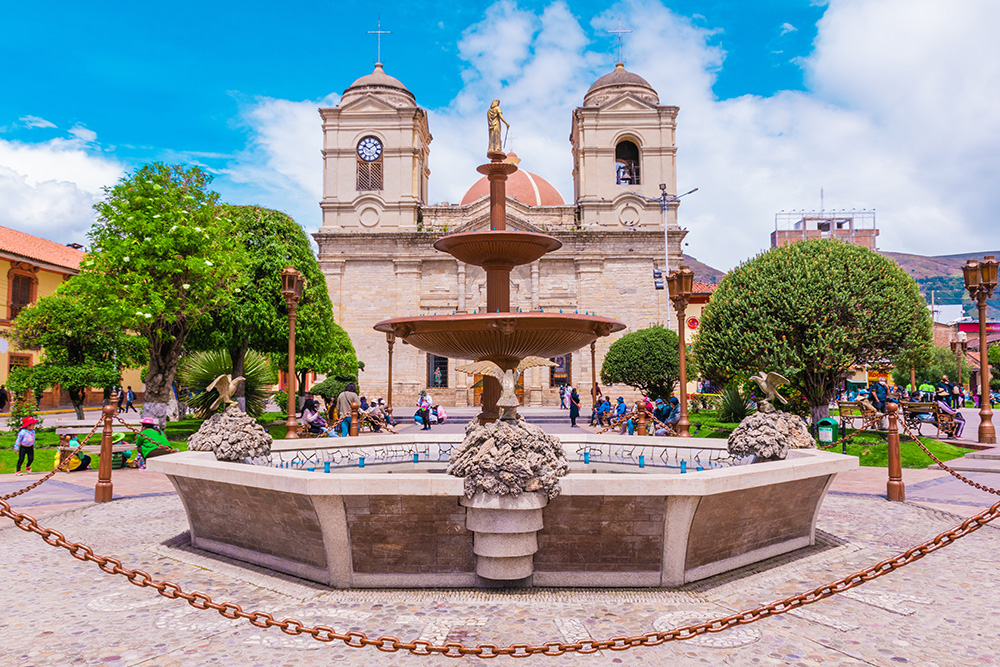 una iglesia con dos torres y frente a su entrada una plaza. En medio de esta una fuente rodeada de unas cadenas que la protegen. A sus costados dos arboles verdes y gente paseando