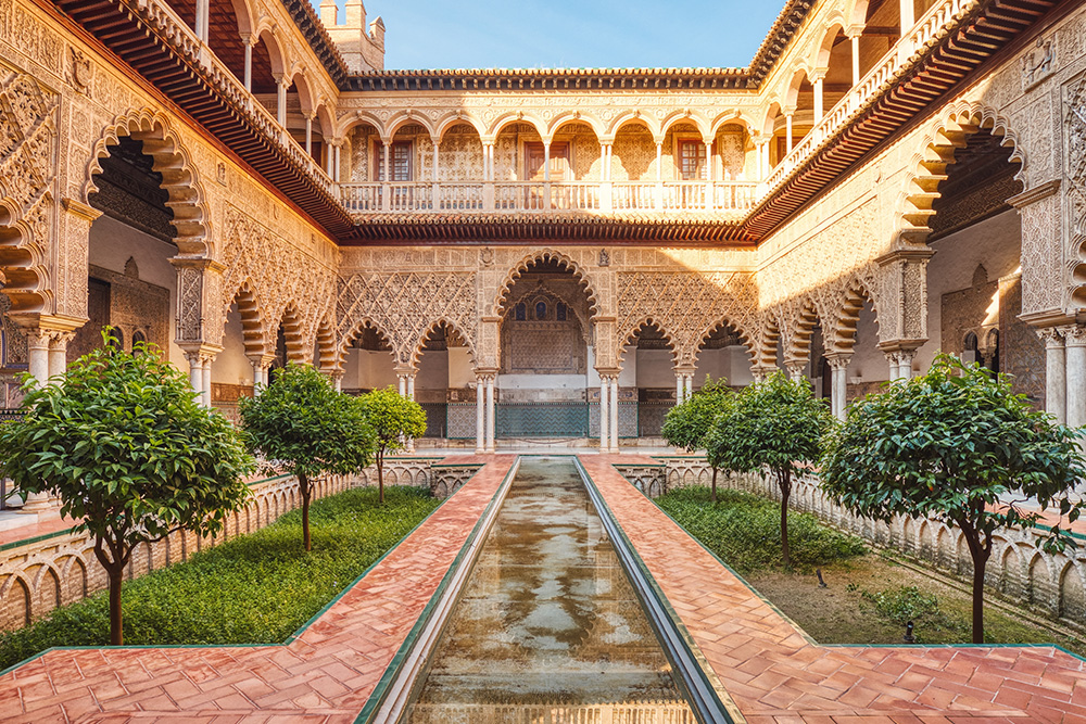 Patio interior de un alcazar con un pasillo en el centro y pequeños arboles en los laterales