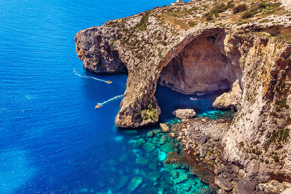 Formación rocosa sobre un mar azul intenso y verdoso. Pequeñas embarcaciones circulas pasando por debajo de la formación rocosa con forma de arco