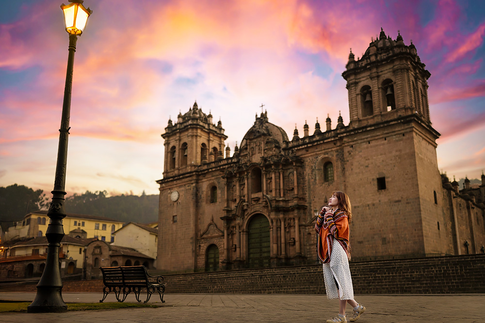 Bajo un cielo rosado se encuentra una iglesia con una escalinata que termina frente a un banco frente a ella y una farola encendida. Mirando la farola una mujer con una capa naranja y unos pantalones blancos