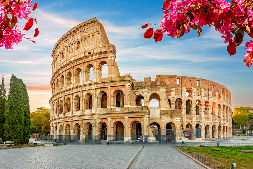 Imagen del Coliseo sin gente alrededor, bajo un cielo azul. En primer plano unas flores rosas