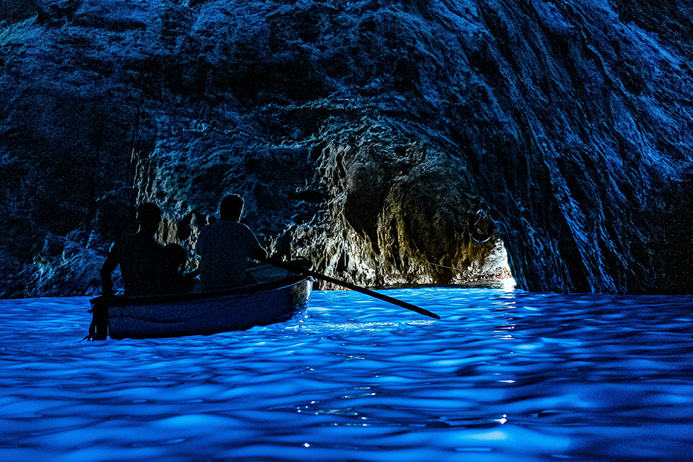 Una pareja en una barca de remos entrando en una cueva maritima. Todo en tonos de azul oscuro 