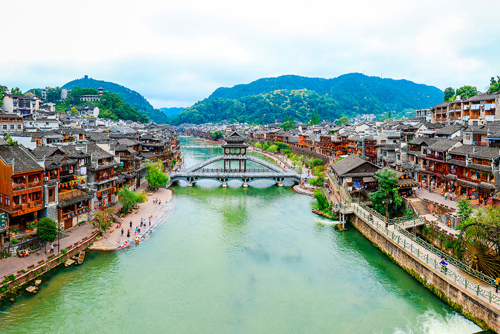 Imagen de un puente cruzando un rio verdoso. A laterales casas en tonos rojos. De fondo montañas
