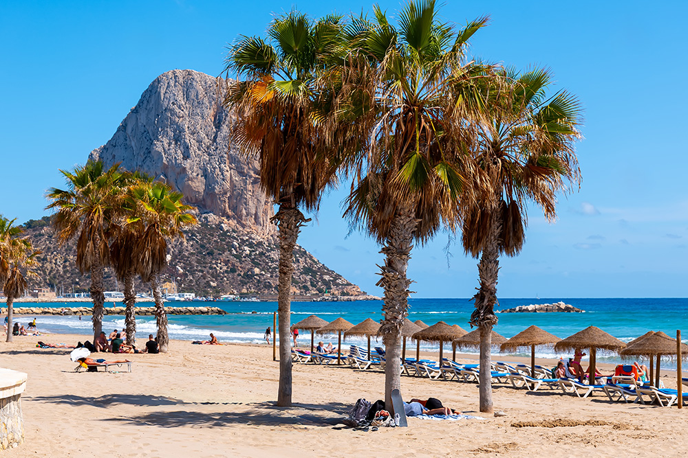 Vista de una playa con un peñon de fondo y en primer plano tres palmeras juntas