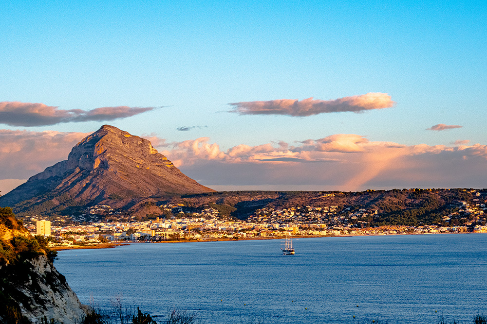 Vista de lejos el puerto con el mar y un pequeño barco en el centro. Montaña al fondo y el atardecer