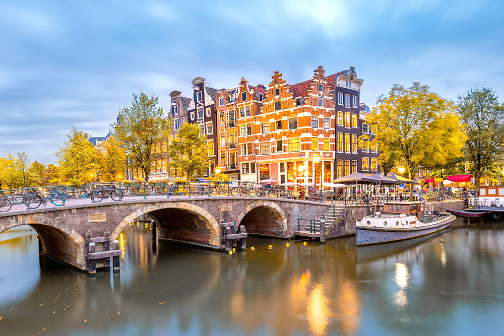 Un canal de Ámsterdam con un puente cruzándolo con bicicletas aparcados. Arboles y edificios clásicos de fondo.
