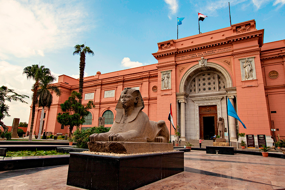 Entrada al Museo presidido por una gran escultura. El edifico en tono naranja claro y rodeado de palmeras