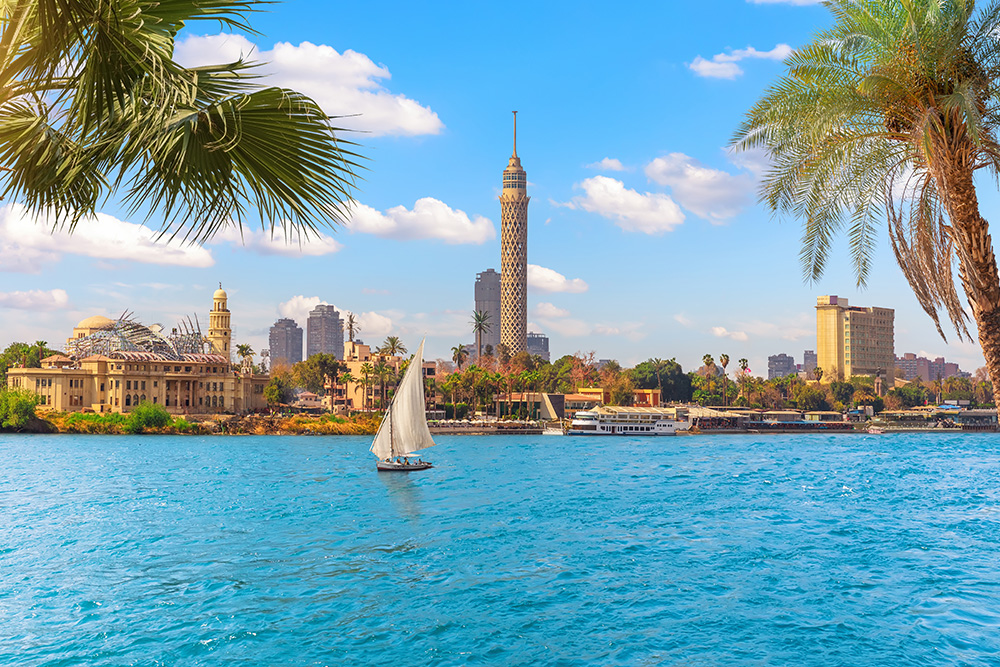 En primer plano el mar en un tono azul claro. Al fondo la ciudad de El Cairo con su torre y las palmeras