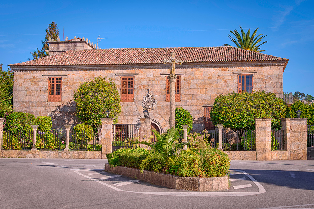 Una casa señorial frente a una carretera. El jardín con muchos arboles verdes y la casa de piedra con tonos marrones
