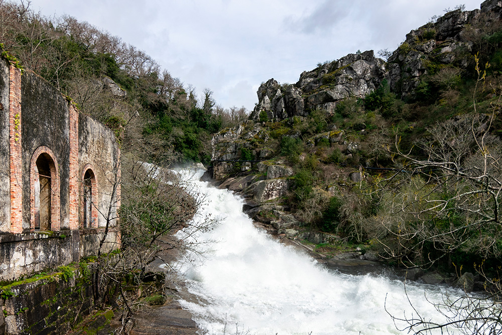 Una cascada entre piedras oscuras y vegetación. A la izquierda un pequeño puente en la piedra