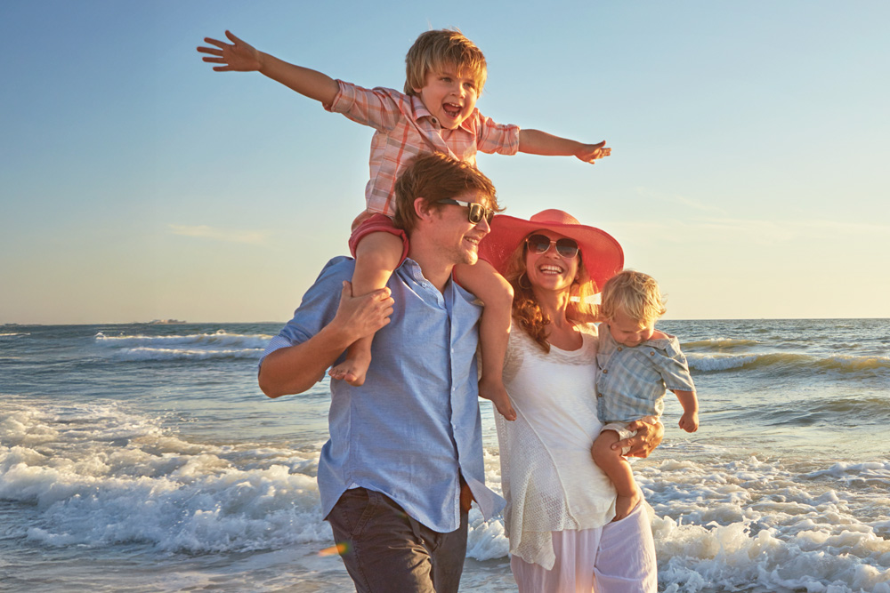 Familia con el niño en hombros del padre y madre con un bebe riendo paseando por la playa 