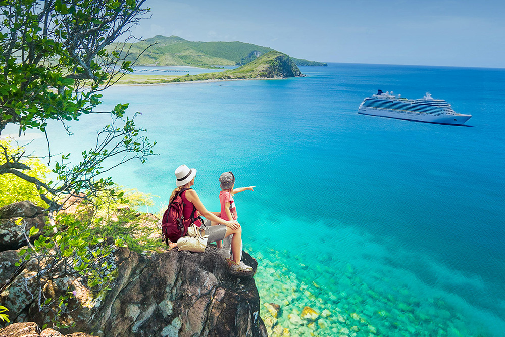 Un padres y su hijo sentados en unas rocas viendo pasar un crucero en un mar azul turquesa 