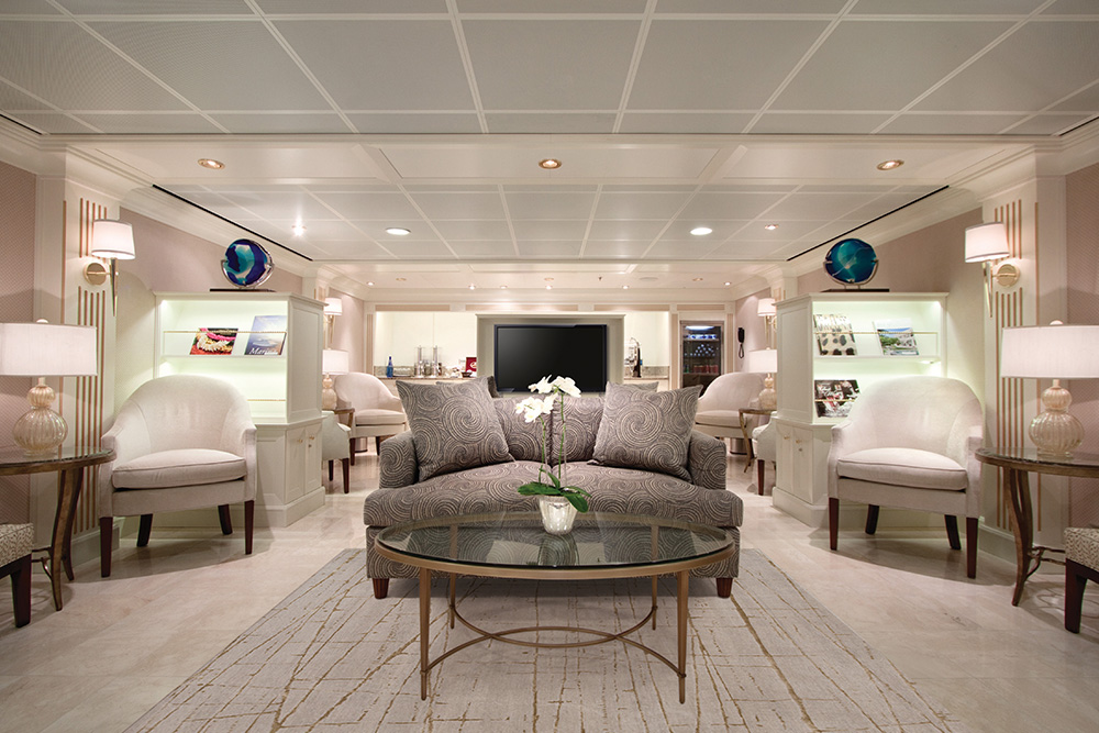 Camarote en tonos neutros. Con un sofa marron en el centro y dos sillones blancos a los lados. De fondo una television