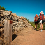 Camino de tierra con pared de piedra al costado. Pareja de espaldas con mochilas caminado 