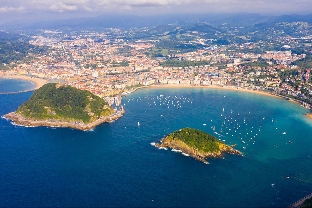 Bahia con una ciudad a la orilla y una pequeña isla en el medio rodeada de veleros 